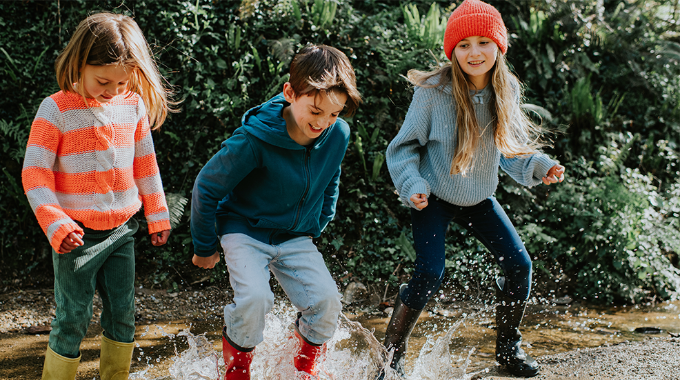 Three young children dressed in bright colours and wellington boots jump in a deep puddle 